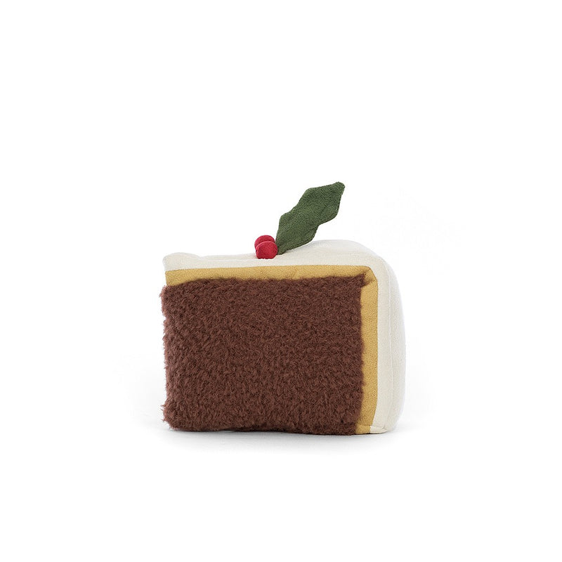 A plush toy resembling a festive slice of Christmas cake, complete with marzipan, icing, and a holly sprig accent. It has twinkling eyes, a stitched smile, and raisin-like boots.