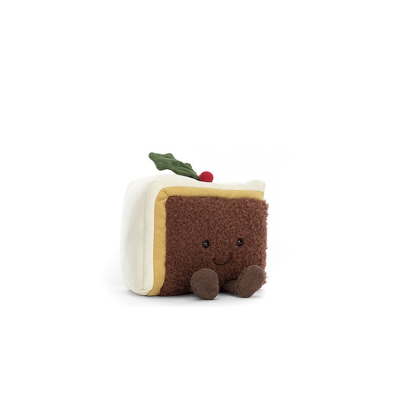 A plush toy resembling a festive slice of Christmas cake, complete with marzipan, icing, and a holly sprig accent. It has twinkling eyes, a stitched smile, and raisin-like boots.