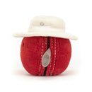 A plush toy resembling a red cricket ball with detailed stitching, wearing a white sunhat.