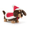 A plush toy of a sausage dog dressed in festive attire, featuring a vibrant red hat and cape with fluffy white trim. The toy has a mix of cocoa and caramel-colored fur, with attentive eyes and floppy ears.